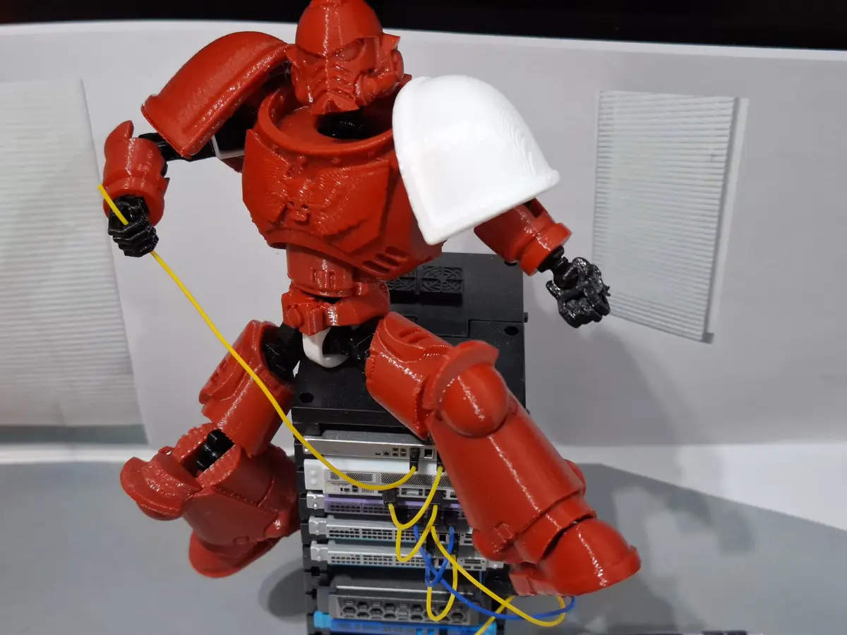A red 3D-printed robot figurine, resembling a Warhammer 40k Space Marine, is posed leaning against a stack of server racks…