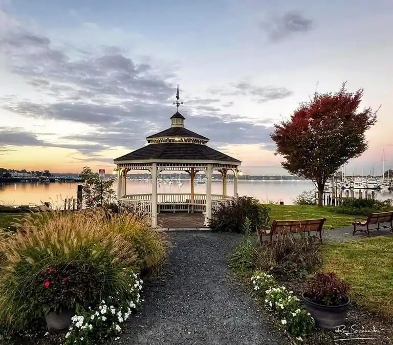 A serene photo of a white wooden gazebo on a waterfront at dusk, with a gravel path leading to it, surrounded by autumn…