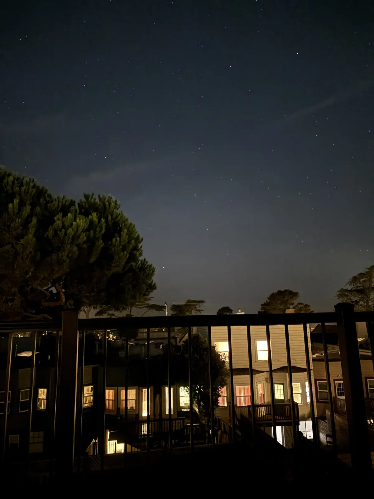 A nighttime photo shows a starry sky above residential houses with lit windows, viewed through a dark railing. The overlaid…