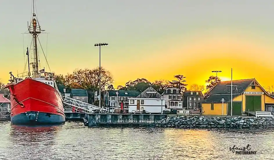 A scenic harbor at sunset with a large red boat docked on the left and a yellow building labeled 'U.S. LIFE SAVING STATION'…