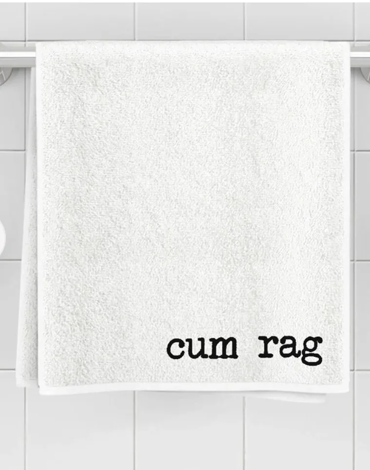 A plain white towel hangs on a silver towel rack against a white tiled bathroom wall. The towel has the words 'cum rag'…