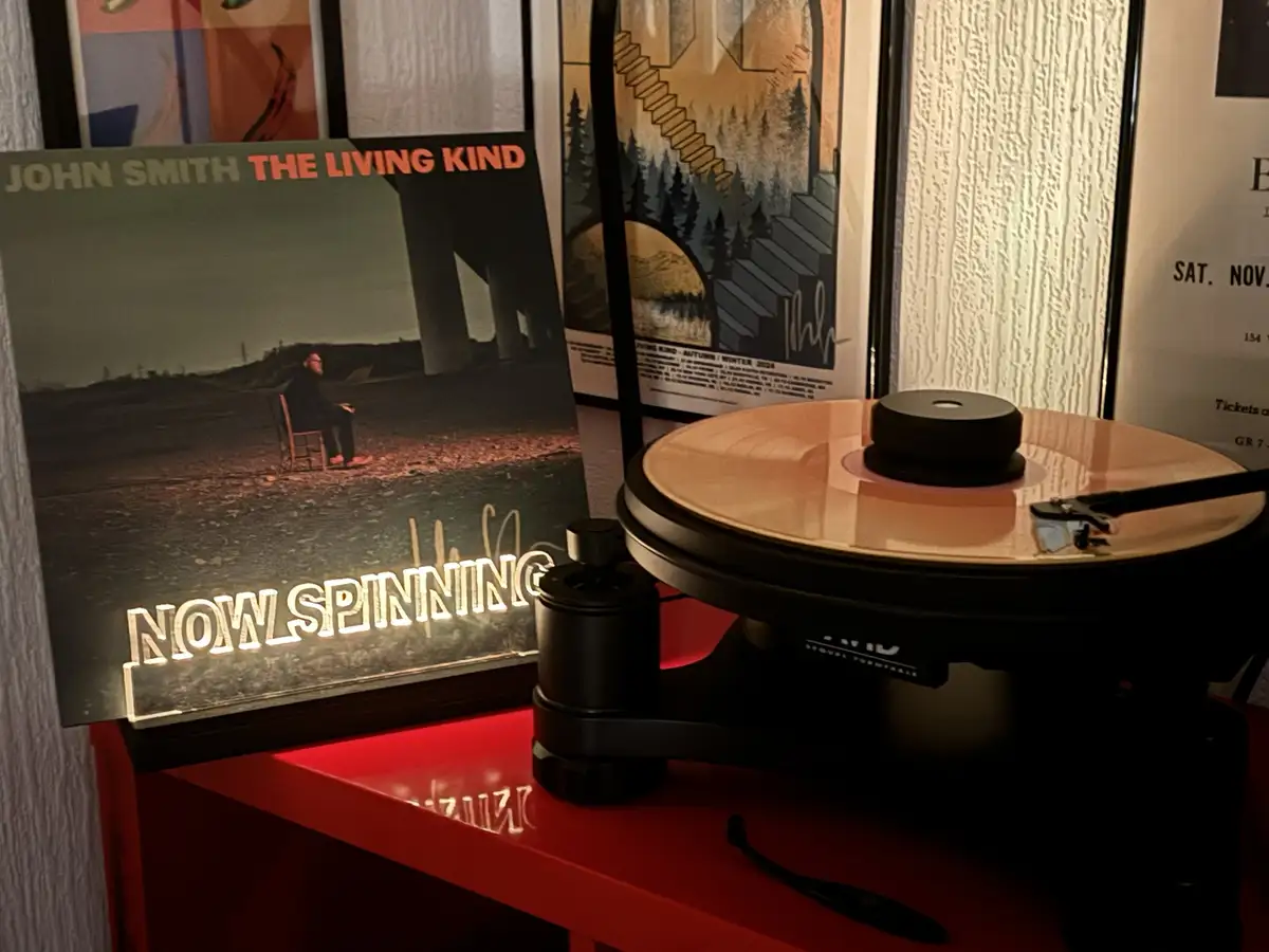 A vinyl record player sits on a red shelf with a record on it, next to a signed album cover for 'John Smith - The Living…