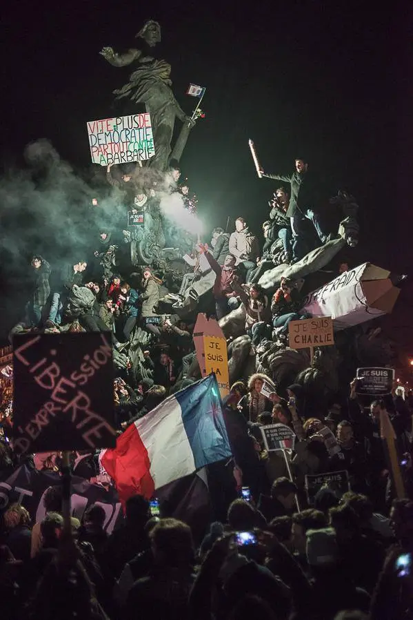 A nighttime protest scene with a large crowd gathered around a statue, holding signs including 'JE SUIS CHARLIE' and…