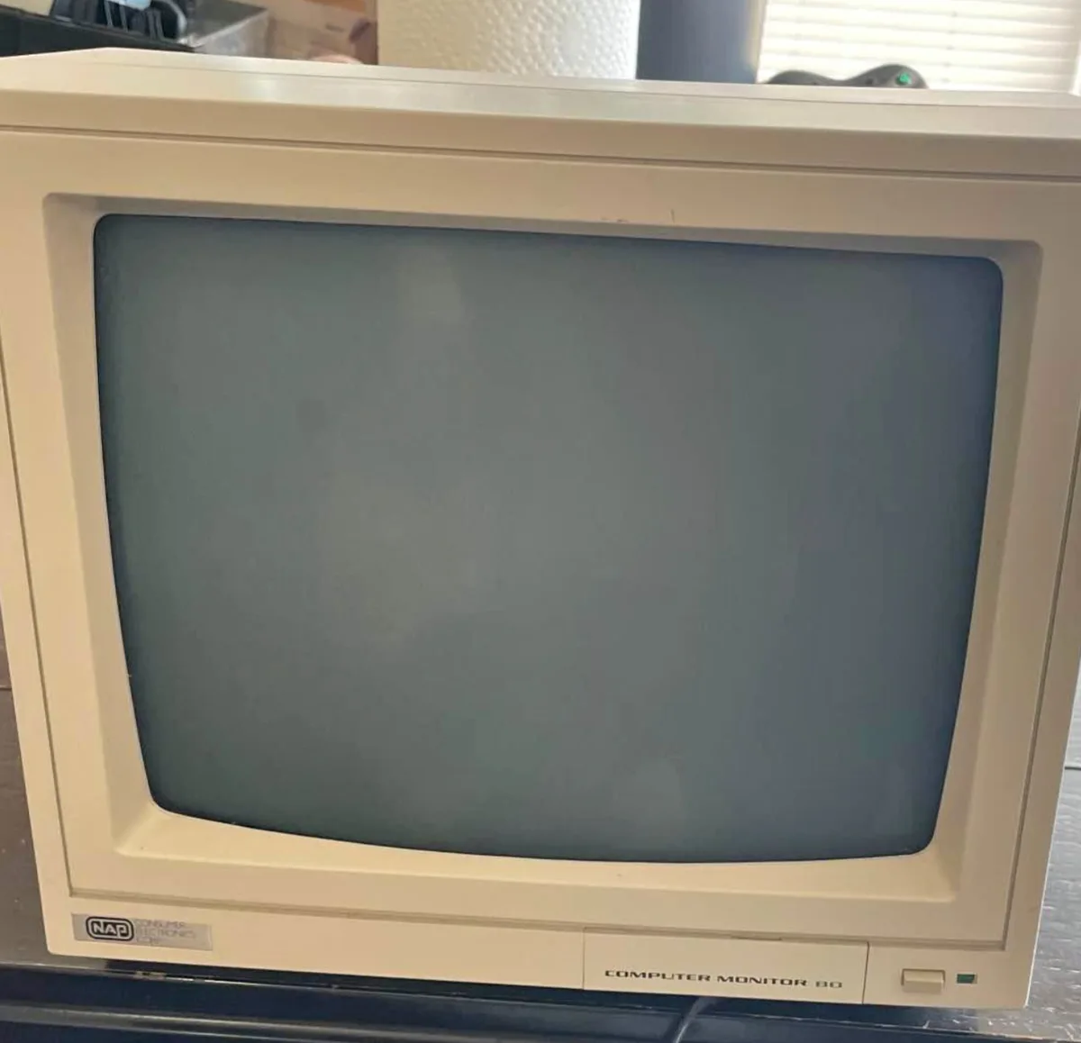 A beige, vintage NAP computer monitor from the 1980s with a blank, dark screen sits on a desk, its label clearly reading…