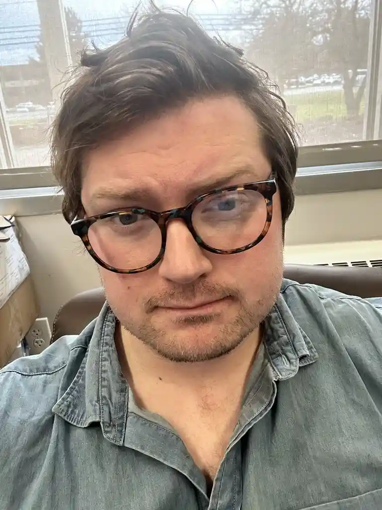 A close-up selfie of a man with brown hair, wearing tortoiseshell glasses and a denim shirt, looking directly at the camera…