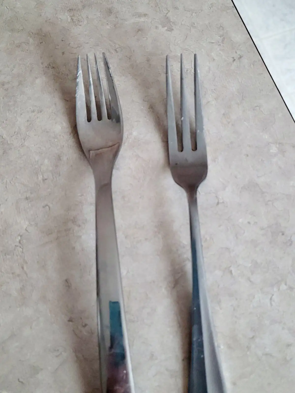Two metal forks lie side by side on a textured beige countertop; the fork on the left has a slightly more worn appearance…