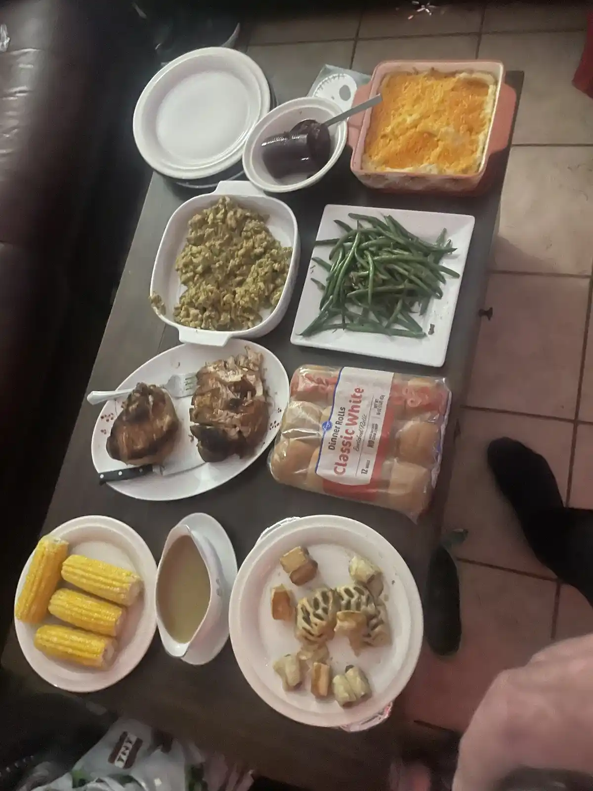An overhead photo shows a coffee table covered with a large, messy spread of Thanksgiving-style food including corn, green…
