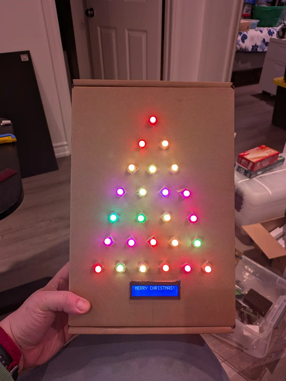 A hand holds a cardboard box decorated with a triangular pattern of colorful LED lights arranged to resemble a Christmas…