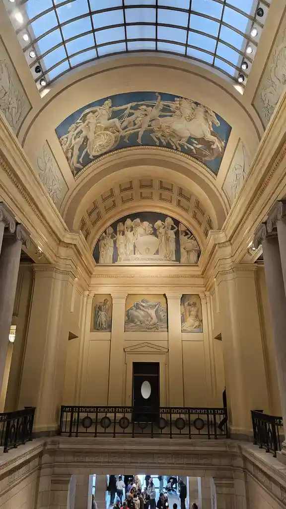 An ornate, grand interior with classical architecture, including large columns, a glass-domed ceiling, and multiple murals…