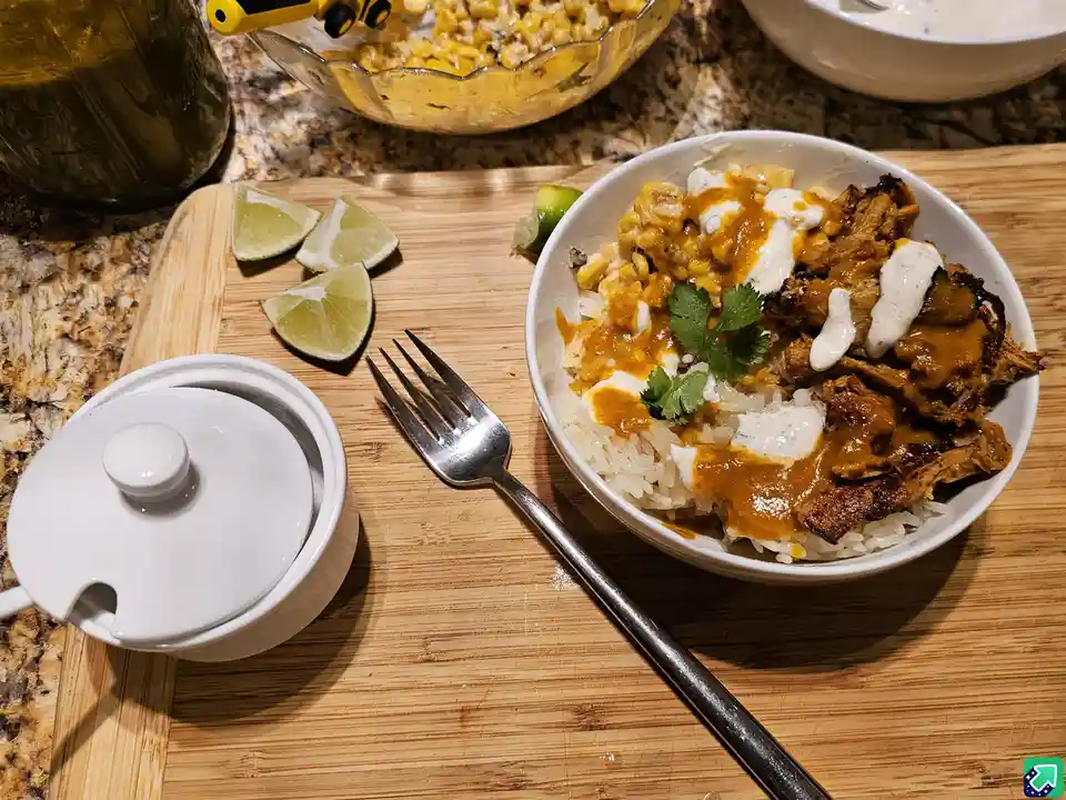 A white bowl of rice topped with shredded meat, corn, orange sauce, and white sauce, garnished with cilantro, sits on a…