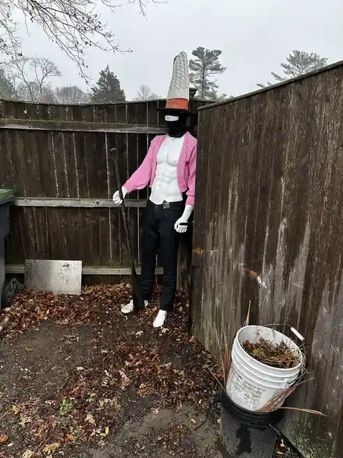 A mannequin stands in a backyard next to a wooden fence, wearing a pink cardigan over a white muscular torso, black pants…
