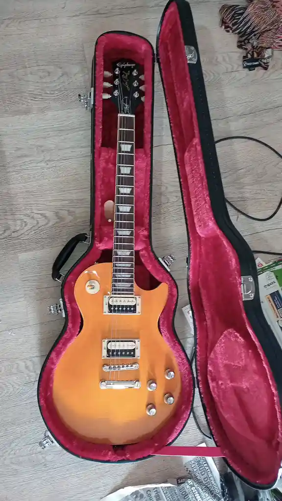 An orange Epiphone electric guitar rests inside an open, red-lined hard case on a light wood floor, with scattered papers…