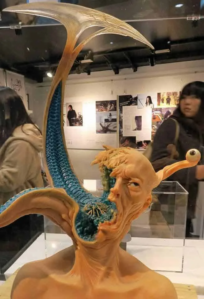 A surreal sculpture of a humanoid figure with a grotesque, open-mouthed expression, revealing blue, textured internal…