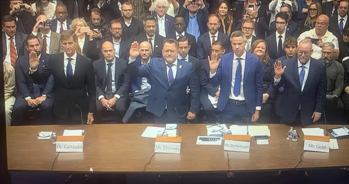 A group of formally dressed men sit at a long conference table with nameplates, some raising their right hands as if taking…