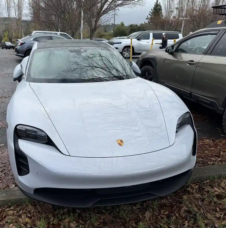 A white Porsche Taycan is parked in a lot with its hood and windshield covered in raindrops and water streaks, with other…