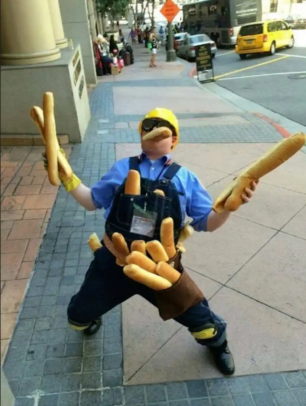 A person dressed as a construction worker with a yellow hard hat, sunglasses, and overalls stands on a city sidewalk in a…