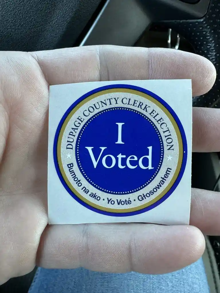 A hand holds a circular blue and gold 'I Voted' sticker from Dupage County Clerk Election, which also includes translations…