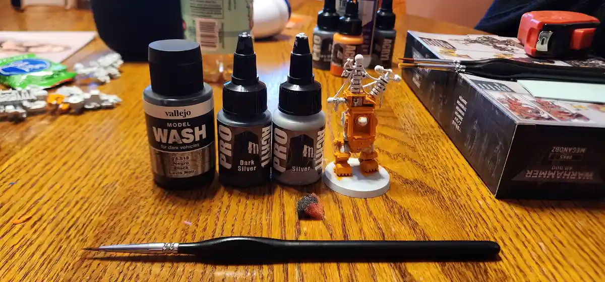 A wooden table holds painting supplies including Vallejo model wash and silver paints, a partially painted orange Warhammer…
