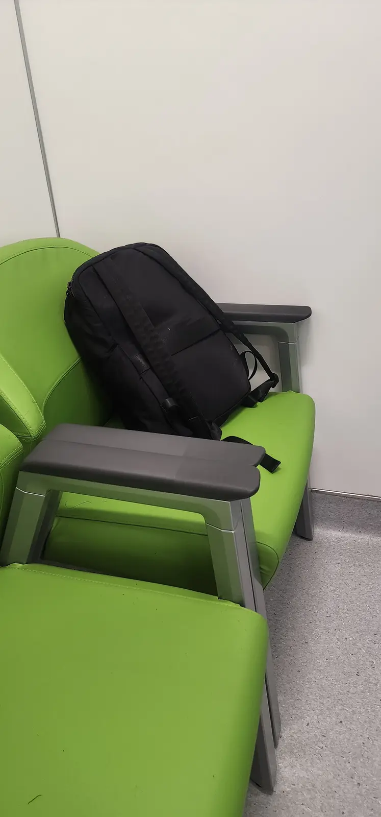 A black backpack is resting on a bright green public seating chair, leaning against the backrest and armrest. The chair has…
