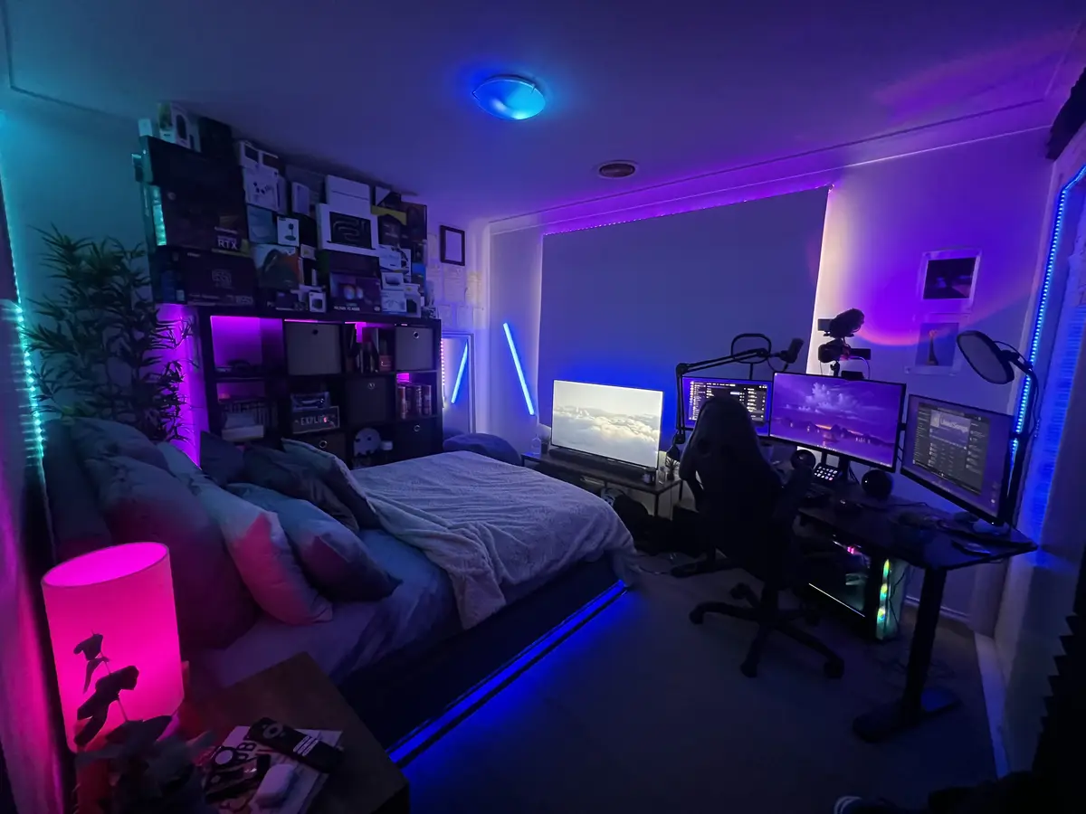 A dimly lit bedroom is transformed into a high-tech gaming and streaming setup, featuring multiple monitors, RGB lighting…