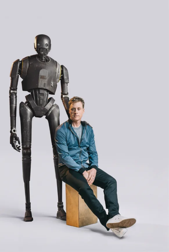 A man in a blue jacket and dark pants sits casually on a wooden block, looking directly at the camera. Standing behind him…