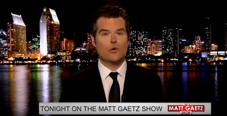 A man in a black suit and tie is shown against a nighttime city skyline reflected in water. A lower third graphic reads…