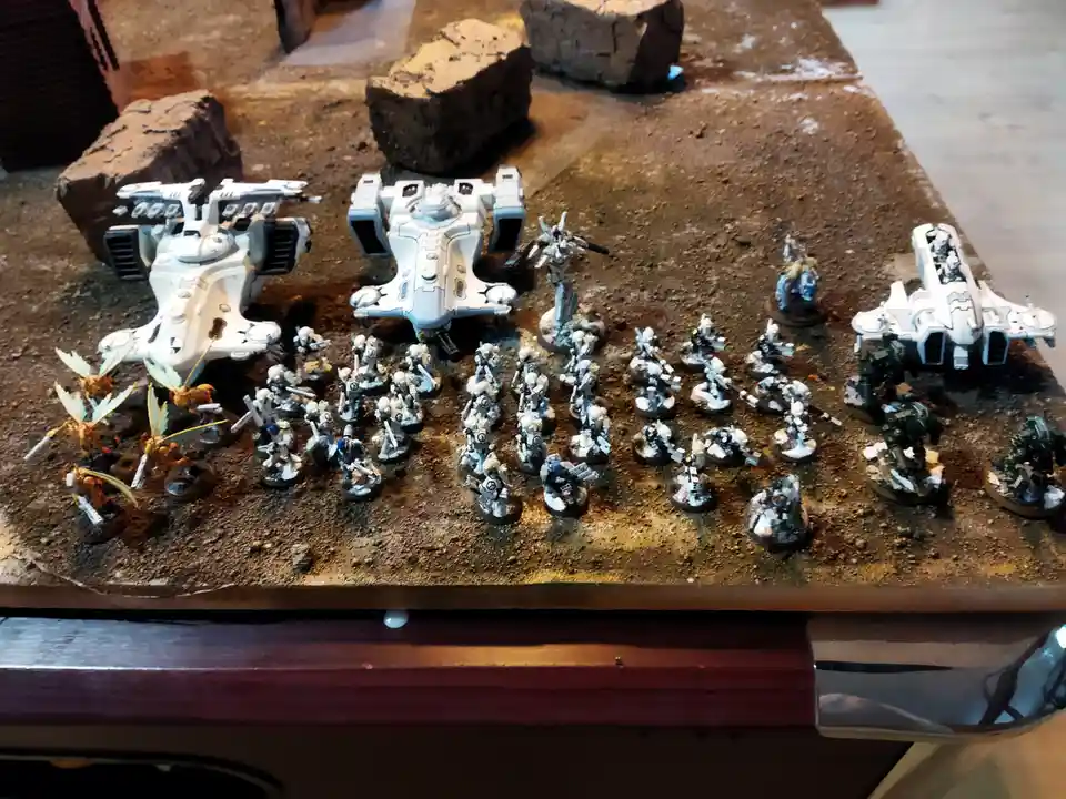 A tabletop wargaming setup shows a large army of white-painted miniature soldiers and vehicles arranged on a brown, rocky…