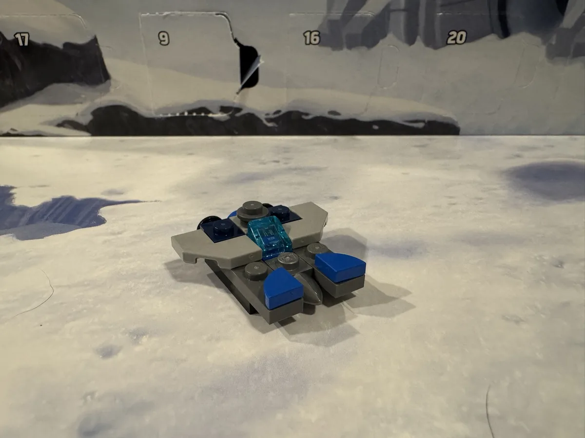 A small, assembled Lego spaceship model with gray, blue, and translucent blue pieces sits on a white, textured surface…