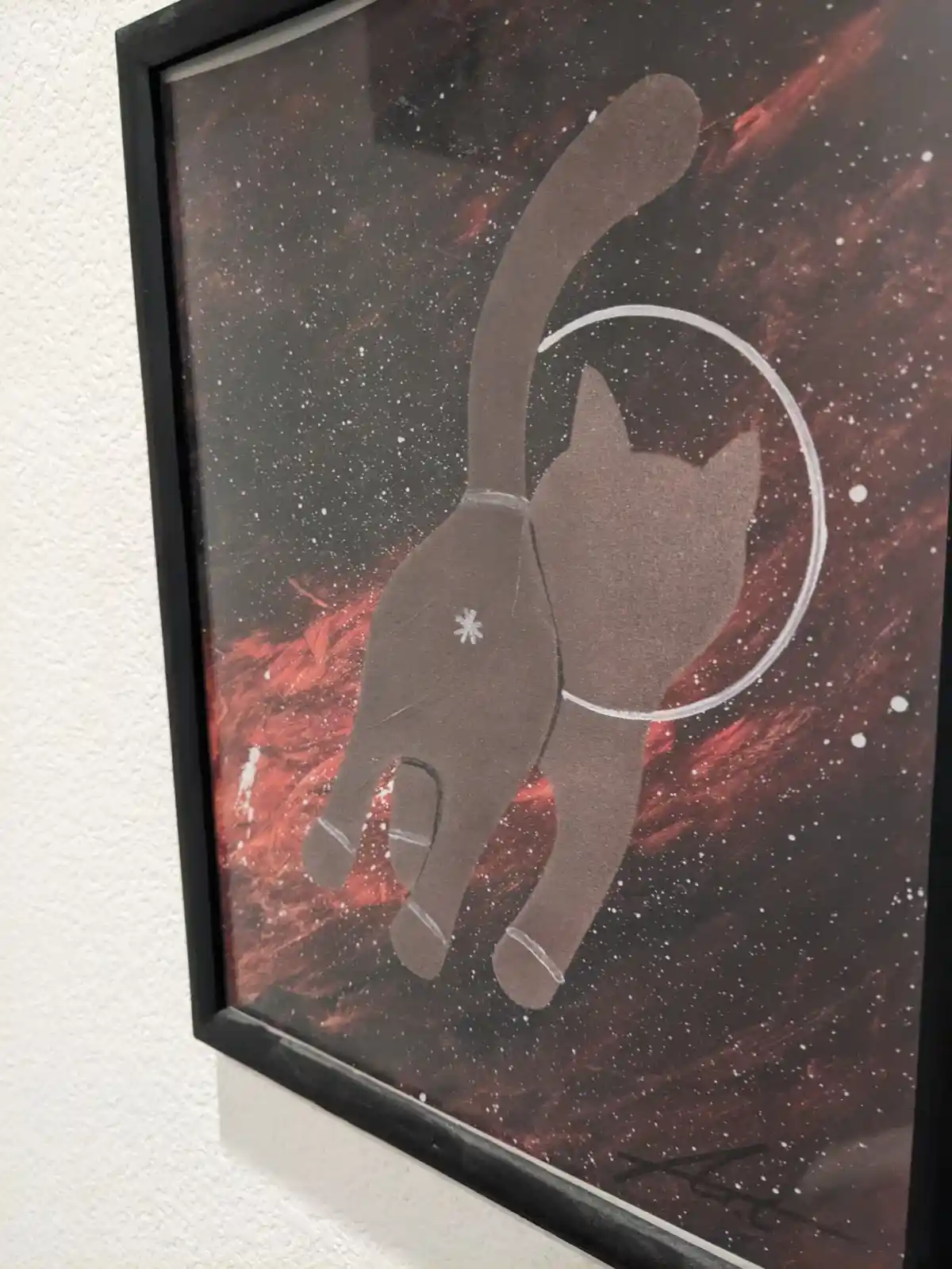 A framed painting shows a brown cat silhouette against a starry, reddish cosmic background. The cat's tail forms a white…