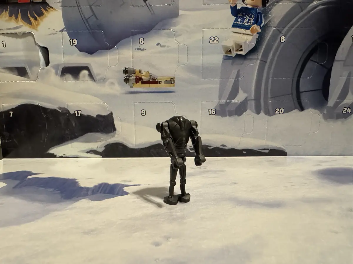 A small black robot figurine stands on a snowy surface in front of a LEGO Star Wars Advent Calendar, which has several…