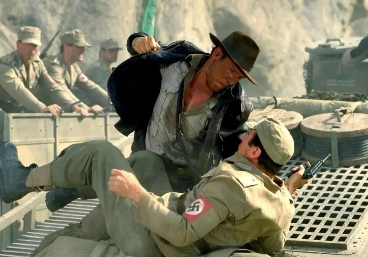 a man in a fedora and vest is shown physically overpowering another man in a military uniform with a swastika armband, while…