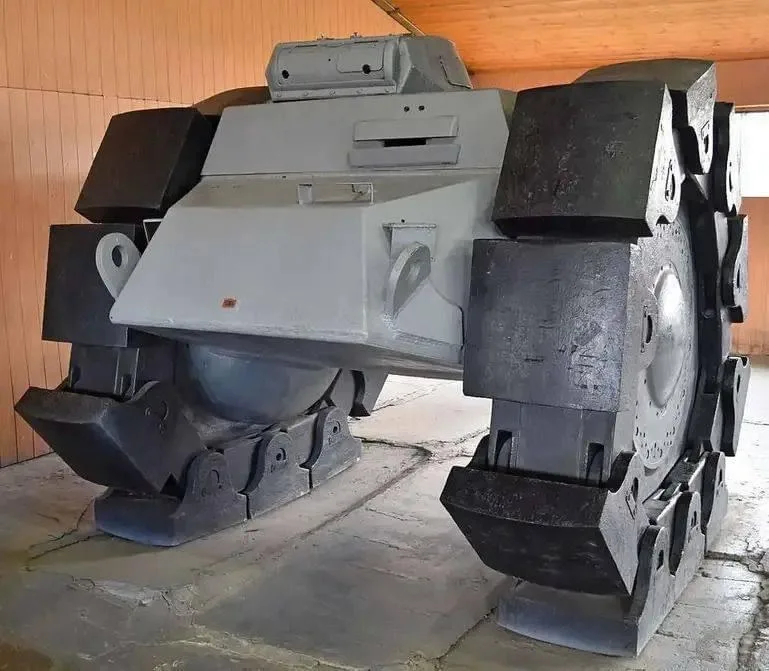 a large, gray, angular military vehicle with heavy treads and a boxy hull is displayed indoors on a stone floor, with wooden…
