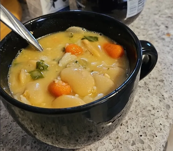 A close-up photo shows a black ceramic bowl filled with a creamy yellow soup containing large white beans, small orange…