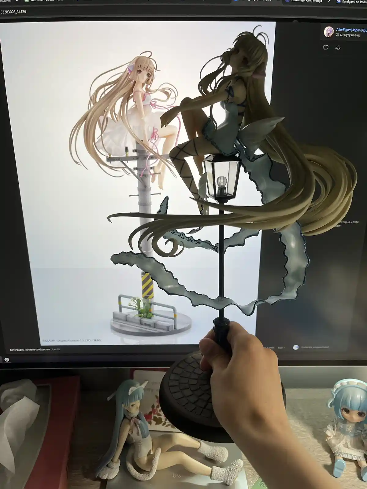 A hand holds a detailed anime figurine of a girl with long blonde hair sitting on a lamppost, positioned in front of a…