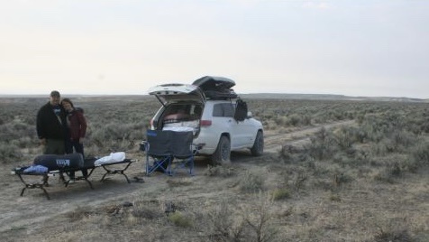 A man and woman stand beside a white SUV parked on a dirt road in a dry, brush-covered landscape. Camping gear, including a…