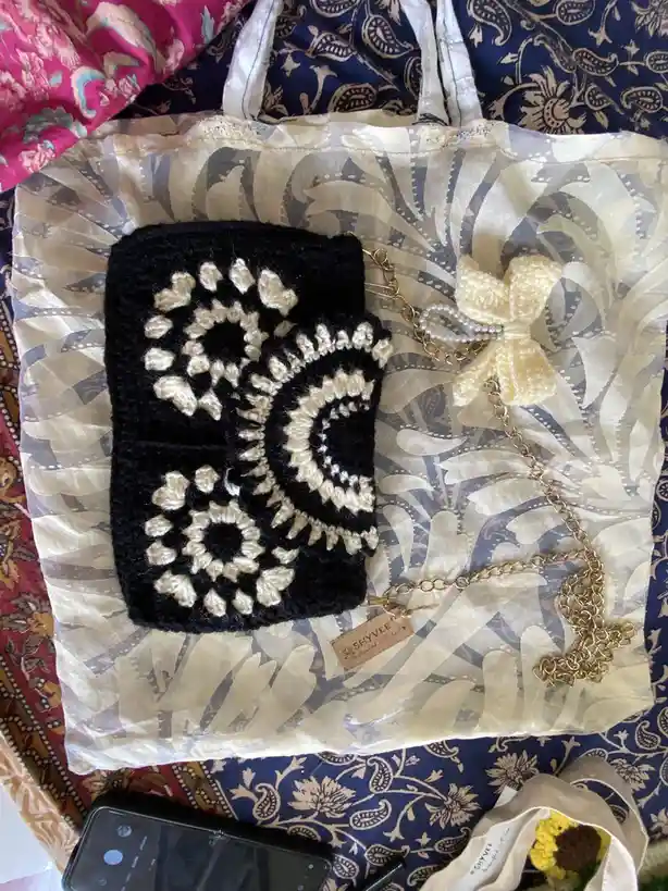 A black and cream crocheted pouch with intricate floral patterns sits on a sheer, patterned fabric inside a tote bag. A gold…