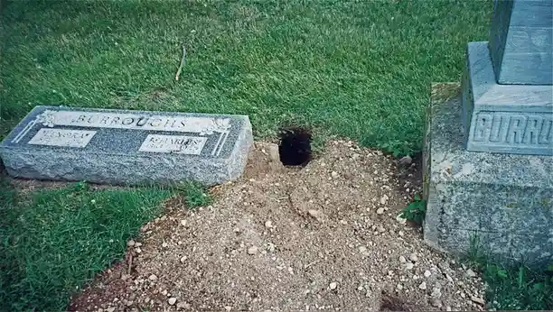 A weathered granite gravestone for the Burroughs family lies in a grassy cemetery, with a large, freshly dug hole in the…