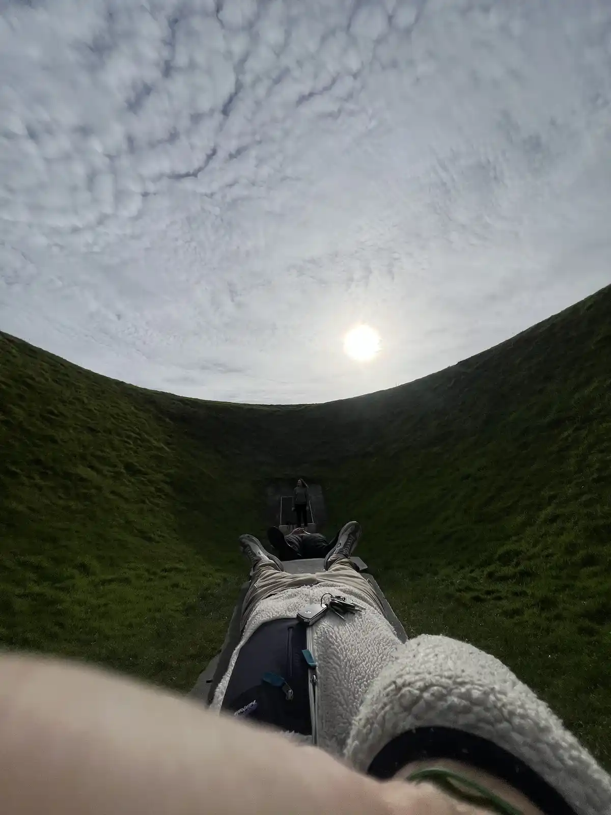 A first-person perspective from someone lying on their back at the bottom of a grassy, bowl-shaped depression, looking up at…