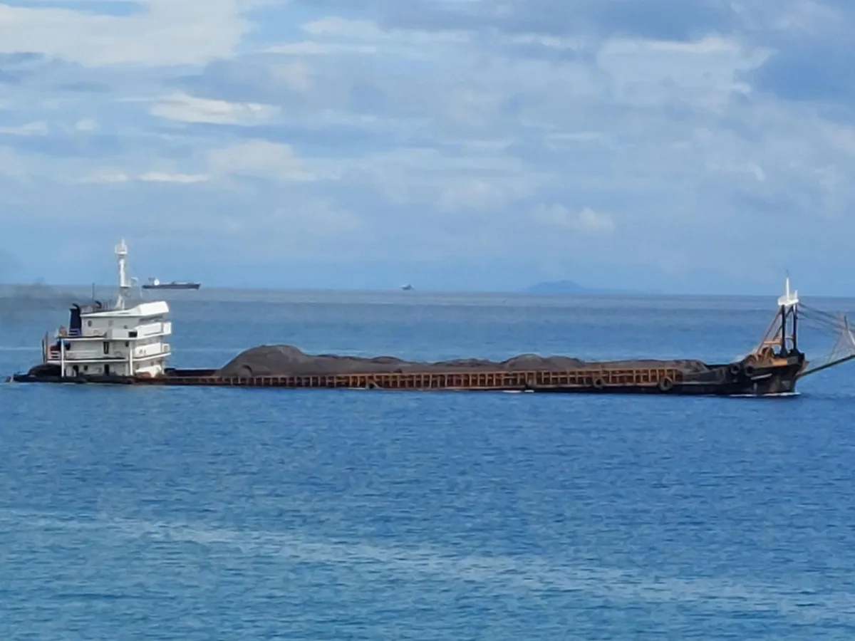 A long, flat-bottomed barge loaded with dark cargo floats on calm blue water under a partly cloudy sky. The barge has a…