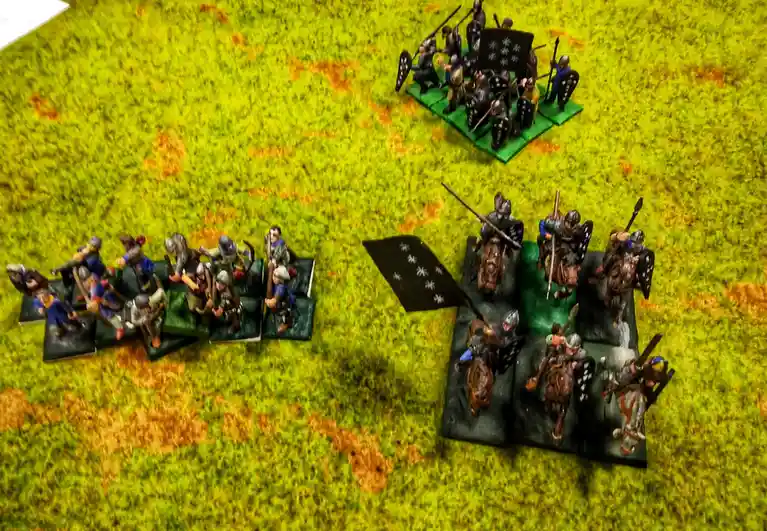 An overhead shot of miniature wargaming figures on a green and brown textured battlefield. Three groups of painted…