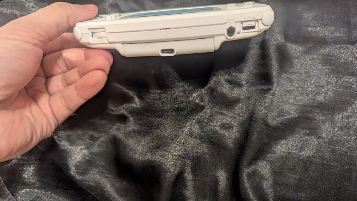 A hand holds a white PlayStation Vita handheld gaming console, showing its side ports and buttons against a dark gray fabric…