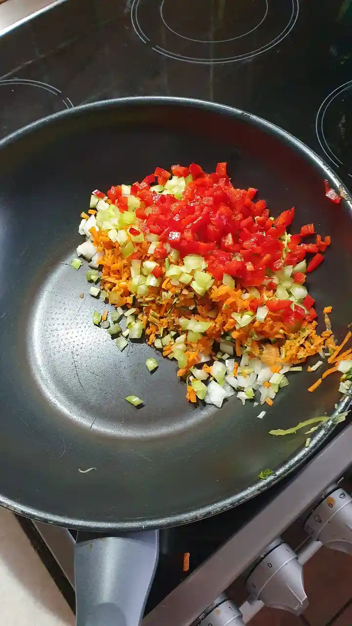 A black frying pan on a stovetop contains a colorful, layered pile of chopped vegetables including red bell pepper, green…