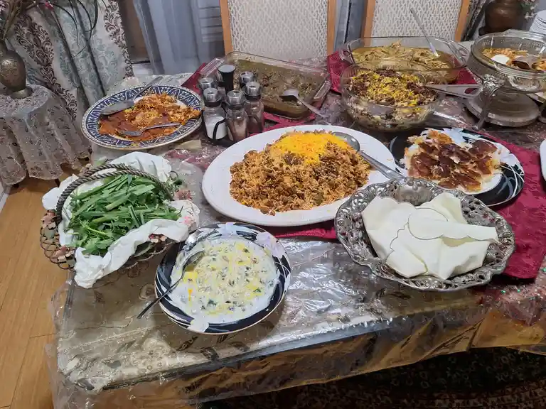 A table is covered with a clear plastic sheet and laden with various dishes, including a large plate of rice with yellow…