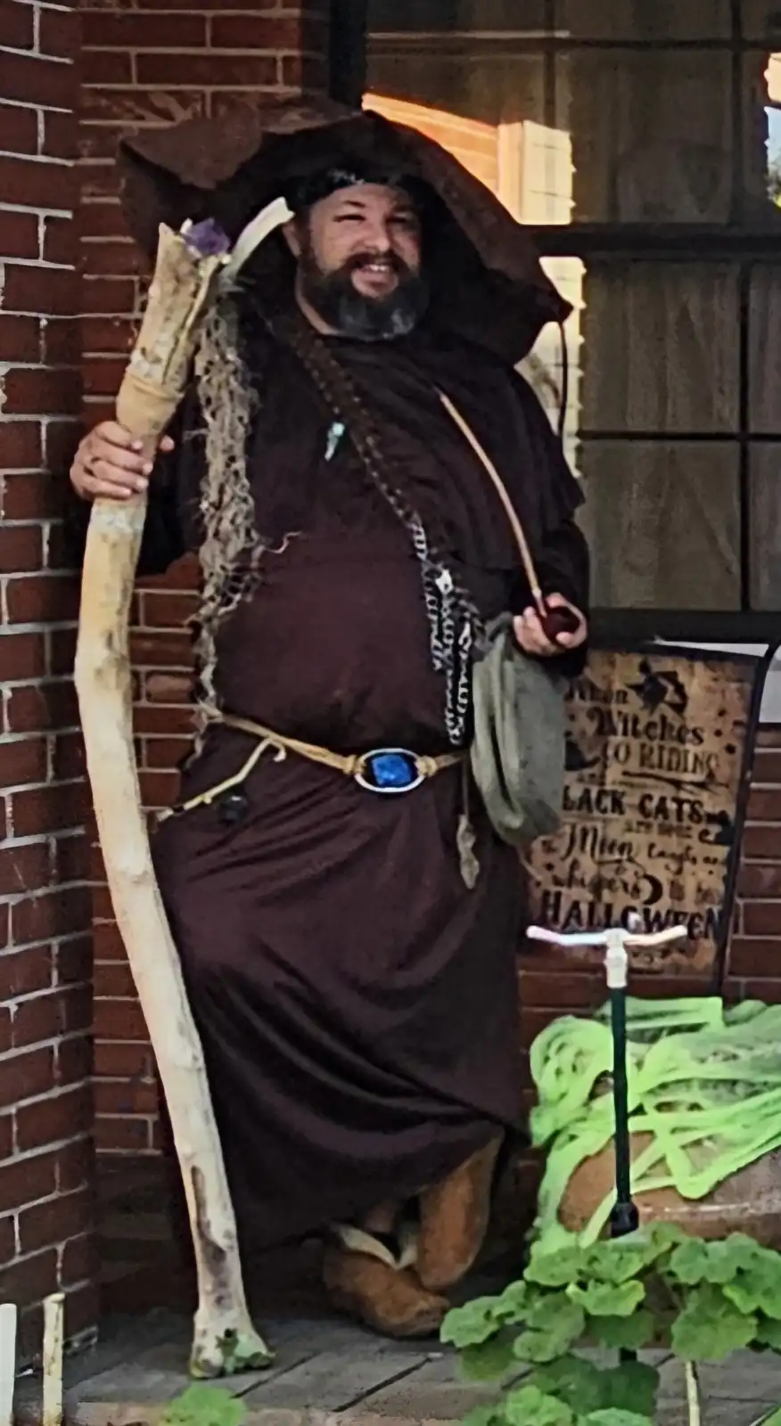 A man dressed as a witch for Halloween, wearing a large, floppy black hat that is visibly worn backwards, a dark robe, and…