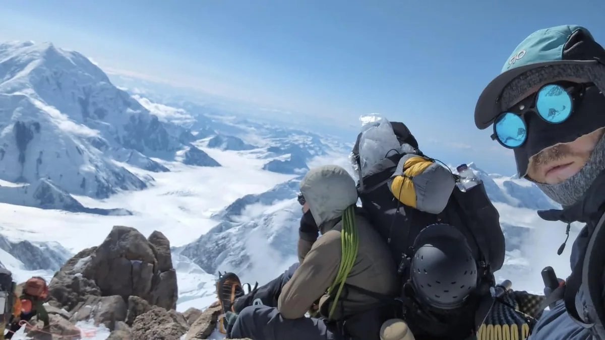 A selfie of a mountaineer at a snowy mountain summit, wearing sunglasses and a face covering, with other climbers in gear…