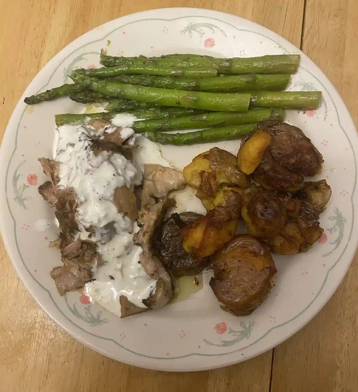 A top-down photo of a white plate with a floral rim, holding roasted asparagus, shredded meat covered in a creamy white…