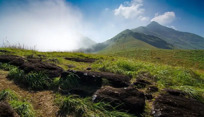 A scenic mountain landscape with green grass, dark rocks in the foreground, and mist partially obscuring distant peaks under…