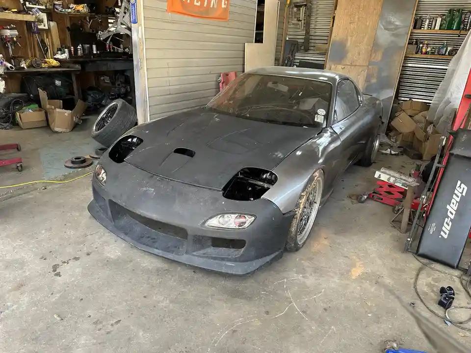 A gray Mazda RX-7 sits in a cluttered garage with its hood and front bumper removed, exposing the engine bay and front…