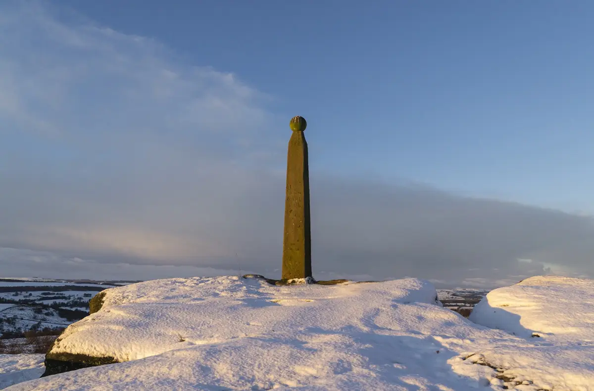A tall, moss-covered stone obelisk stands alone on a snow-covered hilltop under a partly cloudy sky, with a vast, wintry…