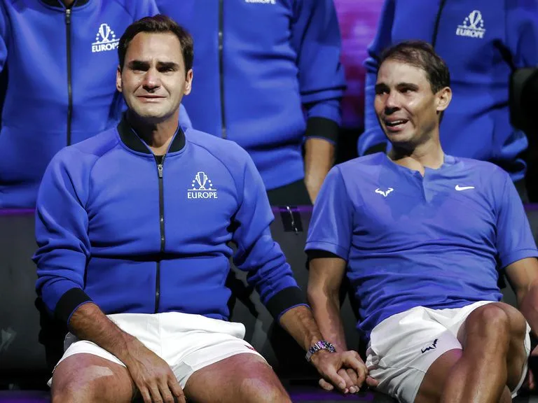 Two male tennis players, Roger Federer and Rafael Nadal, sit side-by-side in blue team attire, holding hands. Federer on the…